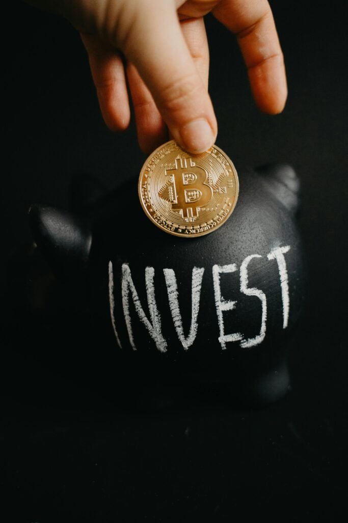 pexels-photo-6781365-6781365 Gold bitcoin inserted into a black piggy bank labeled 'Invest', symbolizing cryptocurrency saving.